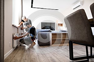 Full length shot of two professional plumbers, workers in uniform fixing or installing heating radiator. Construction