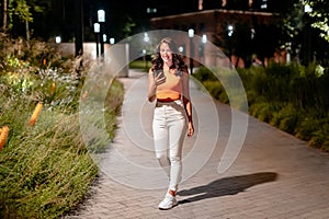 Full length shot of woman walking on the city street at night and using cellphone, texting or browsing online maps app