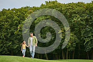 Full length shot of young father and his little daughter holding each other hands while walking in the beautiful green