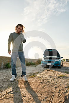 Full length shot of young man looking frustrated, talking on the phone with car service, assistance or tow truck while