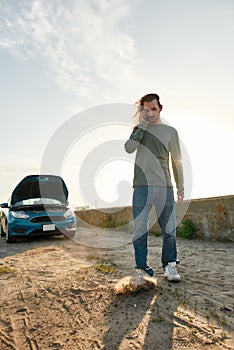 Full length shot of young man talking on the phone with car service, assistance or tow truck while standing near his