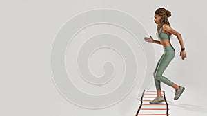 Full length shot of young sportive mixed race woman in sportswear training on agility ladder drill isolated over grey