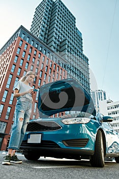 Full length shot of young woman holding phone, calling emergency car service while standing near her broken car with