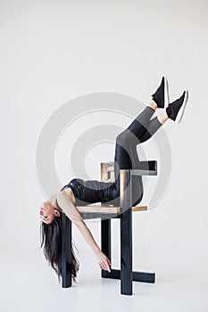 Full-length side portrait of the athletic young woman in sportsuit stretching with the legs on the chair back at the