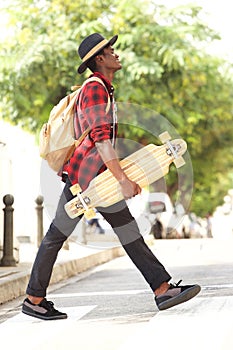 Side portrait of cool young african guy in hat walking outdoors with skateboard and bag