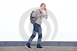 Happy young man walking with bag and talking on mobile phone against white wall