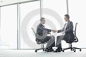 Full-length side view of businessmen shaking hands while sitting on office chairs by window