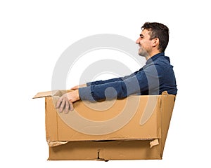 Full length side view of joyful childish man sitting inside a cardboard box isolated over white background