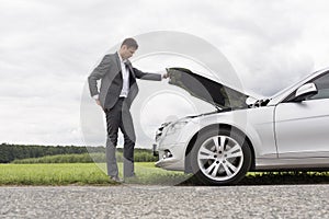 Full length side view of young businessman examining broken down car engine at countryside