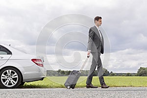 Full length side view of young businessman with suitcase leaving broken down car at countryside