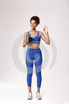 Full length of smiling african-american fitness woman looking healthy and fit, showing okay sign, drinking water during