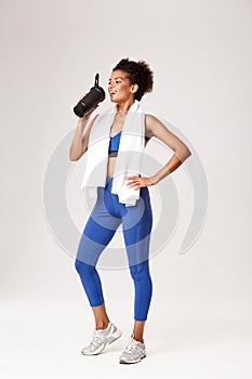 Full length of smiling female athelte in blue sport outfit, drinking protein or water from bottle, standing against