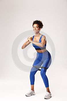 Full length of smiling fit woman enjoying fitness, wearing blue sport outfit, doing squats with resistance band