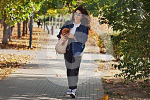 Full length stylish curly brunette woman walks in park and holds dog in bag. Stroll with pet in city