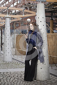Full-length stylish woman model stands at a column and holds a mobile phone