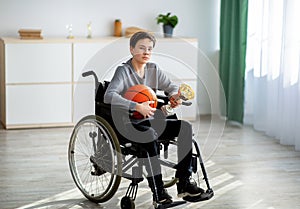 Full length of teen basketball player in wheelchair holding ball and trophy, feeling sad over his injury at home