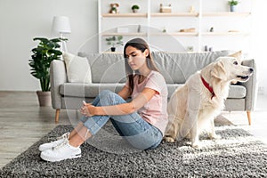 Full length of unhappy young woman sitting on floor with her cute pet dog, feeling lonely and sad at home