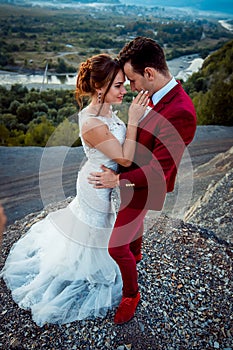 Full-length vertical portrait of the charming newlywed couple standing on the edge of the mountains at the background of