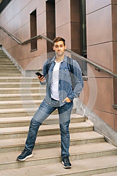 Full length vertical portrait of handsome delivery man with large thermo backpack holding using mobile phone standing on