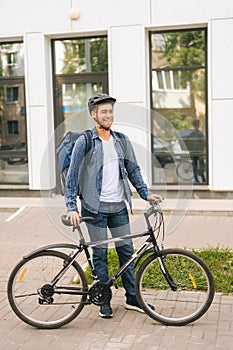 Full length vertical portrait of laughing handsome young delivery man in helmet posing standing near bicycle in city