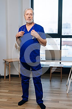 Full length vertical portrait of skilled mature adult male doctor in medical uniform standing in hospital office on