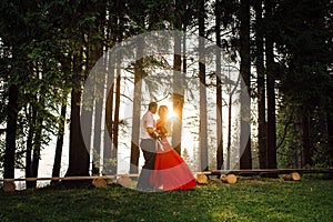 Full-length view of the elegant couple tenderly hugging in the forest during the sunset. The girl in long red drrress is