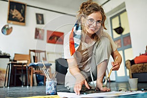 Full-length view of a pretty female artist sitting on the floor in the art studio and painting on paper. A woman painter with