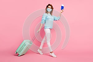 Full length view of pretty wavy-haired girl walking carrying luggage passport wear mask isolated over pink pastel color