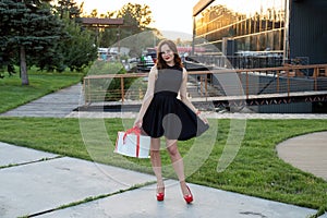 Full length view of pretty young woman with brown hair in little black dress holding white round box with birthday cake wrapped