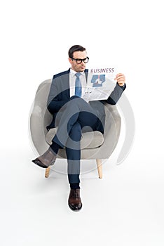 full length view of serious businessman in eyeglasses reading newspaper while sitting in armchair