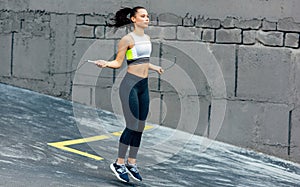 Full-length view of young determined woman skipping with a rope outdoors next to the concrete wall. Fitness female exercising with