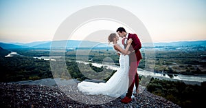 Full-length wedding shot of the adorable newlyweds hugging at the adge of the mountains at the background of the photo