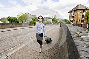 Full length of young asian businesswoman with luggage while answering cell phone walking on sidewalk