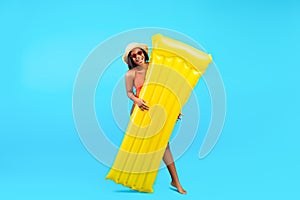 Full length of young black lady holding inflatable lilo and smiling at camera on blue studio background