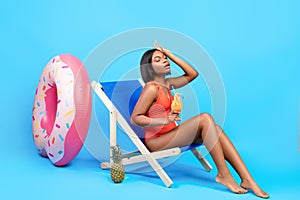 Full length of young black lady suffering from summer heat, holding tropical cocktail while sitting in lounge chair