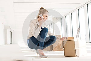 Full length of young businesswoman crouching while using mobile phone and laptop in new office