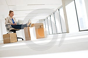 Full length of young businesswoman using laptop with feet up on moving box in office