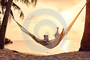 Full length of young woman freelancer celebrating success, keeping arms raised while lying in hammock on the tropical beach at