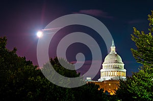 Full moon behind United States capitol building illuminates it marble dome at night with trees silhouetted in foreground