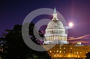 Full moon and clouds stand in sky behind marble dome of United States capitol building at night with trees silhouetted in national