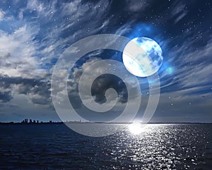 Full moon on cloudy fluffy dark night starry sky at sea and moonlight reflection on water on horizon city silhouette nature landsc