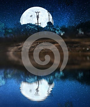 Full moon with milky way stars hidden behind the mountain and trees at the night time on sea with lunar reflection on the water.