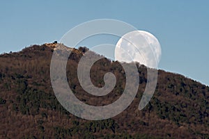 The full moon sets early in the morning behind a mountain against the blue sky