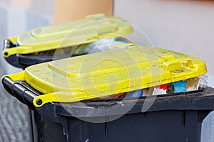 Full recycling plastic yellow packing bins with yellow lids against an urban backdrop, signaling waste management