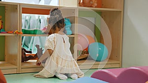 full shot of a little pretty girl sitting on the floor and playing with toys on shelves, kindergarten concept