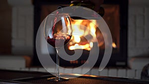 Full shot pouring splashing red wine from bottle into glass goblet fireplace in background