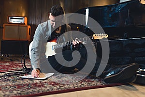 full shot of a young guitarist man sitting on the floor with a bass guitar and writing notes in the studio