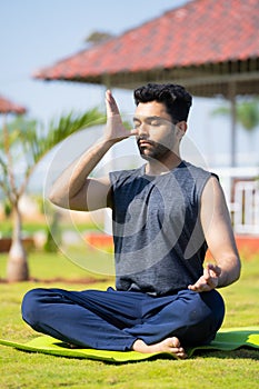 full shot of Young man doing nostril breathing exercise or pranayama yoga by eyes closed at park - concept of zen