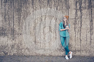 Full size of cheerful young lady, standing near concrete wall outside, smiling, with crossed legs and plastic cup with coffee, in