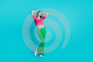 Full size photo of adorable gorgeous girl wear pink knit top green trousers hold arms behind head isolated on blue color